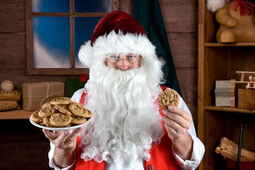 Santa Claus in his workshop holding a plate full of fresh baked Chocolate Chip cookies in one hand and a single cookie in the other. 