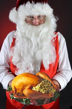Santa Claus serving a fresh Roasted Thanksgiving or Christmas Turkey with all the trimmings. He is holding the platter in both hands in front of his torso. Vertical on a light to dark red background.