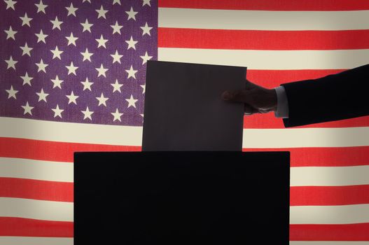 Voting Concept. Closeup of a man’s hand, in silhouette,  placing a ballot into a box in front of an American Flag. 