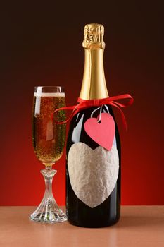 Closeup of a champagne bottle decorated for Valentines Day. A glass of champagne is next to the bottle. The bottle has a red ribbon and heart shaped tag and a blank heart shaped label.