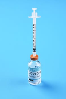 Closeup of a Covid-19 vaccine vial with a syring sicking straight up out of the bottle on a blue background.