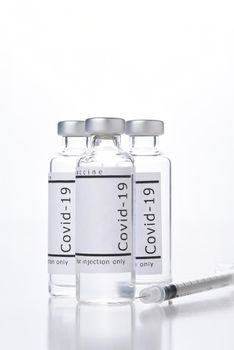 Three vials of Covid-19 Vaccine and syringe on white with reflection.