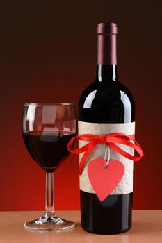 Closeup of a wine bottle decorated for Valentines Day. A glass of wine is next to the bottle. The bottle has a red ribbon and heart shaped tag and a blank label.