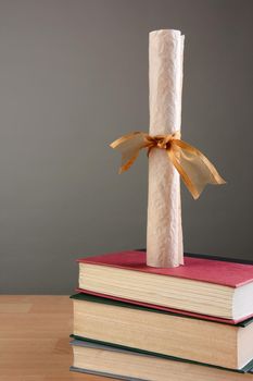Closeup of a parchment diploma standing on a stack of school books, against a light to dark gray background. Vertical format with copy space.