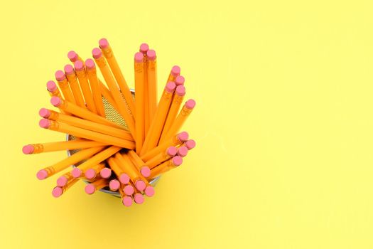 Looking down on a pencil cup filled with new pencils. On a bright yellow background with the eraser end  towards the camera. Horizontal Format with copy space.