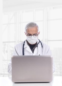 Middle aged doctor wearing a surgical mask sitting at a desk using a computer in a modern medical facility. Vertical format. 