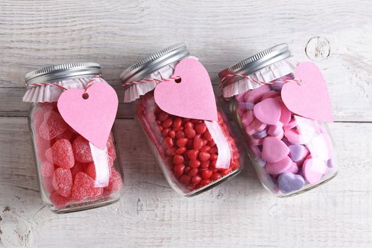 Canning jars laying on their sides filled with candy hearts and for Valentine's Day on a rustic wood table. The jars have blank heart shaped gift tags hanging from the neck.