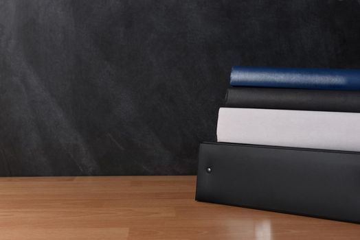 A binder with textbooks on a teachers desk in front of a chalkboard.