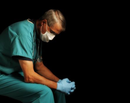 Exhausted Middle Aged doctor in scrubs, gloves and mask sitting with head down amid the Covid-19 crisis, profile portrait against black.