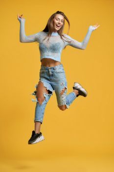 Front view of cheerful girl in full height with long brown hair raising left leg up in jump wearing short blue top with long sleeves, ripped jeans.