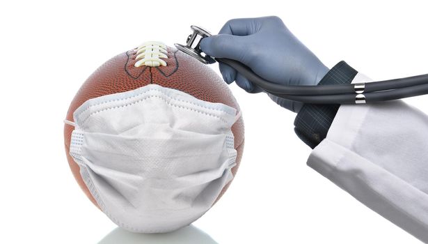 Covid-19 and Sports Concept. An American Football With Surgical Mask and a doctors hand holding a stethoscope to the ball to check its condition. 