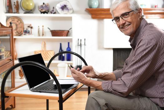 Mature man working from home with his laptop and cell phone. Man is looking at the samera ans smiling.