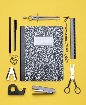 Office and back to school supplies on a yellow background. Looking down on the all black and chrome tools from an overhead angle. The items are arranged in a rectangle forming a frame.
