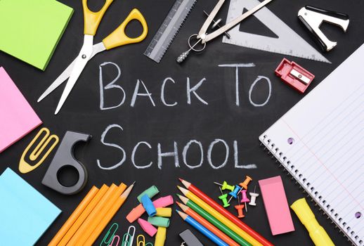 Back to school written on a chalkboard. The words are surrounded by school supplies including, paper, scissors, pencils, erasers, paper clips, compass, ruler, push pins and other necessities for students.