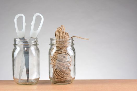 Closeup of two jars with string and scissors for wrapping packages. Horizontal format on a wood desk top against a light to dark gray background with copy space.
