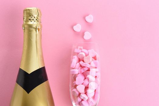 Closeup of a bottle of champagne and a flute filled with pink candy hearts for Valentines Day. On a pink background with copy space.