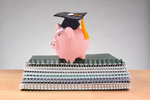 Closeup of a stack of spiral notebooks on a teachers desk with a piggy bank wearing a graduation cap, representing the high cost of education.