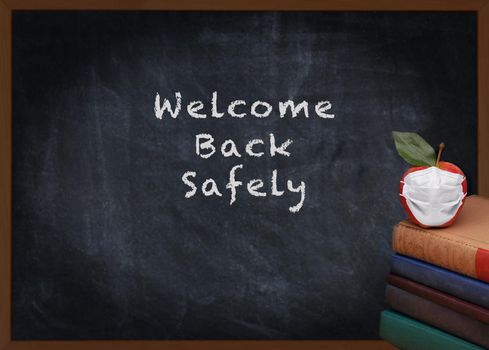 Back to School Concept. A stack of books and a red apple with a Covid-19 Mask on top in front of a chalkboard with Welcome Back Safely.