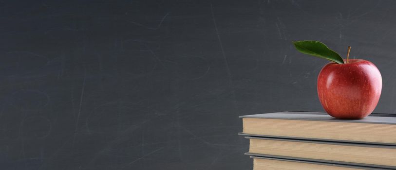 Back to school concept: Text books with a red apple in front of a blank chalk board. Banner sized image with copy space.