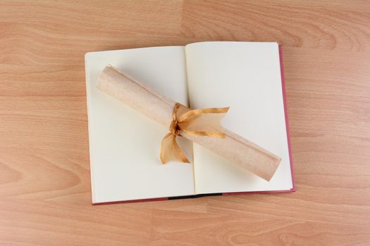 HIgh angle view of an open school book with a parchment diploma rolled and tied with a gold ribbon laying across the blank pages. Horizontal format on a wood desk.