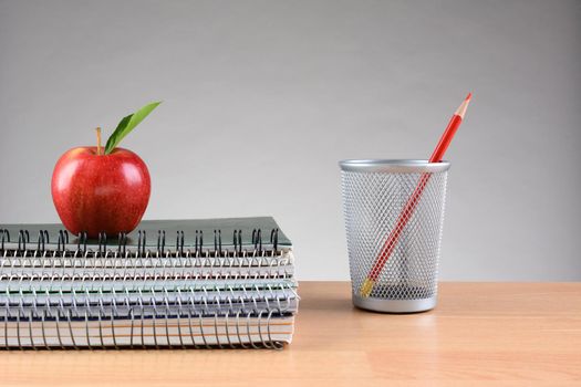 Closeup of a stack of notebooks with and apple on top and a pencil cup with a solo red pencil on a teachers desk. Horizontal format with copy space on a light to dark gray background.