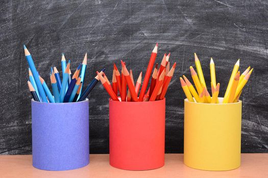 Closeup of colored pencils in matching pencil cups in front of a school room chalkboard. The cups are covered in red, blue and yellow construction paper and filled with the same color pencil.