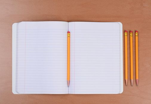 An open theme book with a pencil in the fold and three more sharpened pencils to the side. Horizontal format on a wood classroom desk.