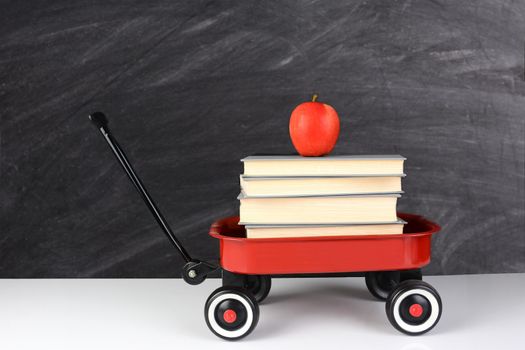 A red wagon full of school books with an apple on top. horizontal format in front of an out of focus chalkboard. Back to School concept with copy space.