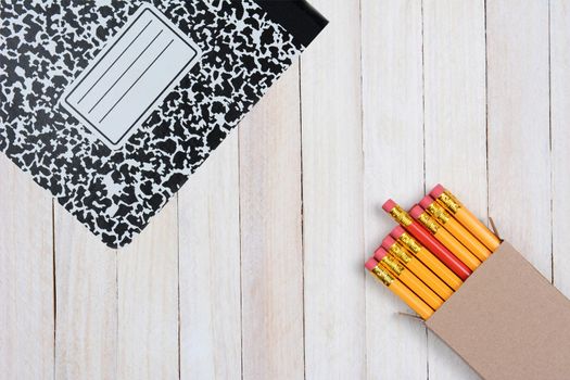 High angle shot of a box of pencils and a composition book on a white wood surface. Objects are in opposite corners of the frame.