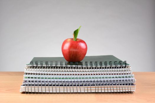 Closeup of a stack of spiral notebooks on a teachers desk with a single red apple centered on top. Back to school concept with copy space on a light to dark gray background.