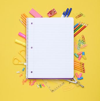 An open spiral notebook laying on assorted school supplies. Back to School concept. Equipment includes, pens, pencils, erasers, compass, scissors, paper clips and more.