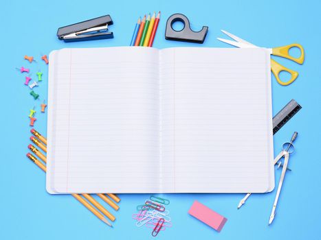 An open notebook surrounded by school supplies. Back to School concept with compass, eraser, stapler, tape dispenser, push pin, pencils, paper clips, scissors and a ruler on a blue background.