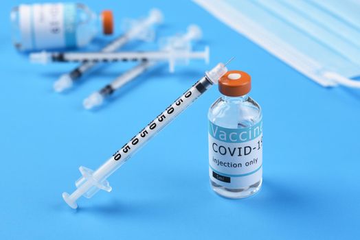 A vial of Covid-19 vaccine with a syringe leaning on the bottle with more syringes, a bottle and surgical mask in the background. 