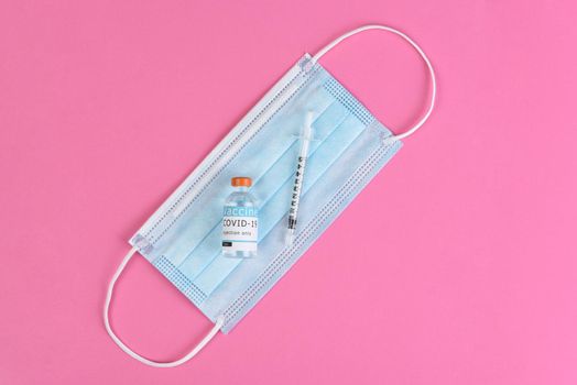 Equipment to administer a Covid19 Vaccination. Flat lay with surgical mask, syringe and vaccine vial, on pink background.
