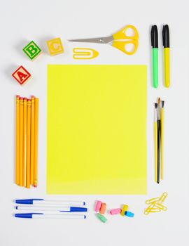 A variety of school supplies arranged around a sheet of blank yellow paper. Vertical format with copy space