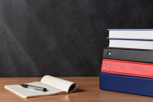 A stack of three different binders on desk in front of black board with two books and note pad and pen.