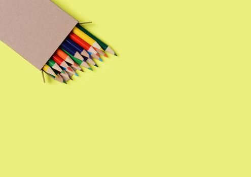High angle shot of a box of multi-colored pencils on a yellow background. The pencils are part way out of the box which is at an angle in the lower left corner leaving copy space.