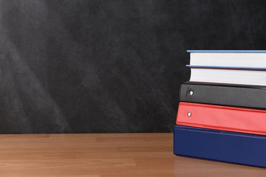A stack of three different binders on desk in front of black board with two books.