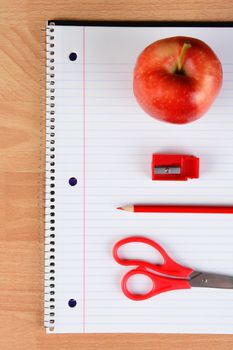 Overhead view of a red pair of scissors, red pencil, sharpener and apple on an open spiral notebook on a wood desk. Back to school concept.
