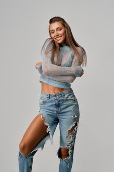 Attractive smiling girl with long brunette hair hugging herself in short blue top with long sleeve and ripped jeans on isolated grey background at studio. Concept of style, fashion.