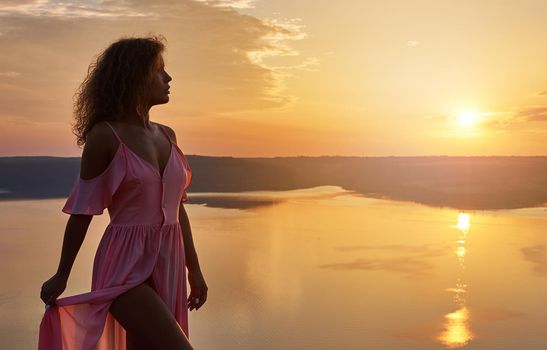 Charming, attractive girl wearing in pink light dress with bare shoulders standing on shore and looking at sky. Beautiful sundown. Lady resting and admiring landscape in evening.