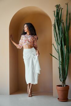 Charming dark-haired woman posing near big green cactus in trendy summer blouse and skirt. Indoors photo of enchanting european girl in high heels standing in decorative arch of the wall.