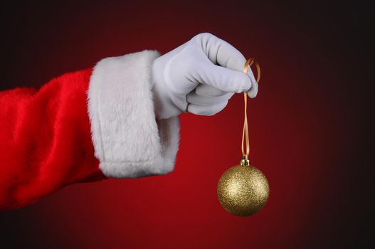 Santa Claus holding a gold sparkly tree ornament over a light to dark red background. Horizontal format showing only hand and arm.