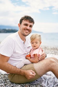 Smiling dad holding a little girl in his arms sitting on a pebble beach. High quality photo