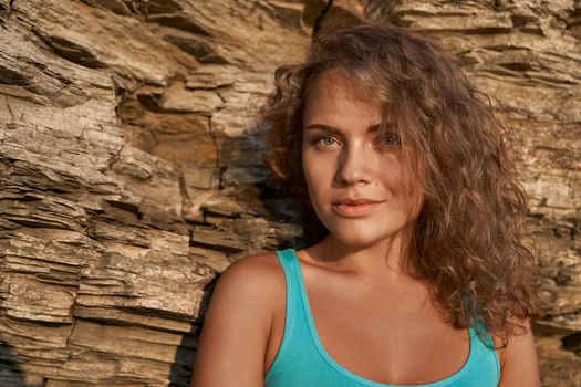 Portrait of beautiful girl with long blonde curly hair, plump lips on background of rocky in sunny day. Attractive model wearing in blue sleeveless t shirt looking at camera.