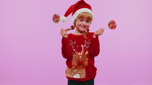 Funny girl in New Year sweater, hat holding candy striped lollipops, hiding behind them, fooling around, dancing isolated on violet studio wall background. Happy Christmas celebration merry holiday