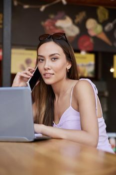 young asian pretty woman in pink dress sitting at diner, using laptop, calling by smartphone. attractive female in light pink dress working at cafe. lifestyle portrait, modern communication