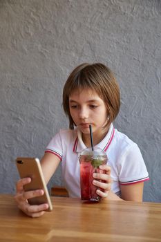 schoolgirl holds smartphone, drinks lemonade through straw. little girl reading information on phone, drinking red icy cocktail at table of cafe. modern communication technology, using digital devices