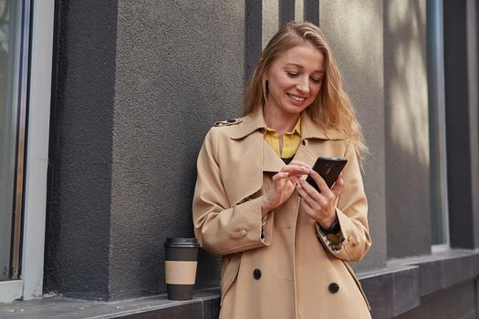caucasian woman in beige trench coat using smartphone outdoors on sunny day, reading news, social media. attractive female street portrait at fall or spring. autumn lifestyle, modern communication