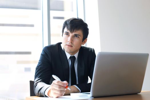 Portrait of happy businessman sitting at office desk, looking at camera, smiling.